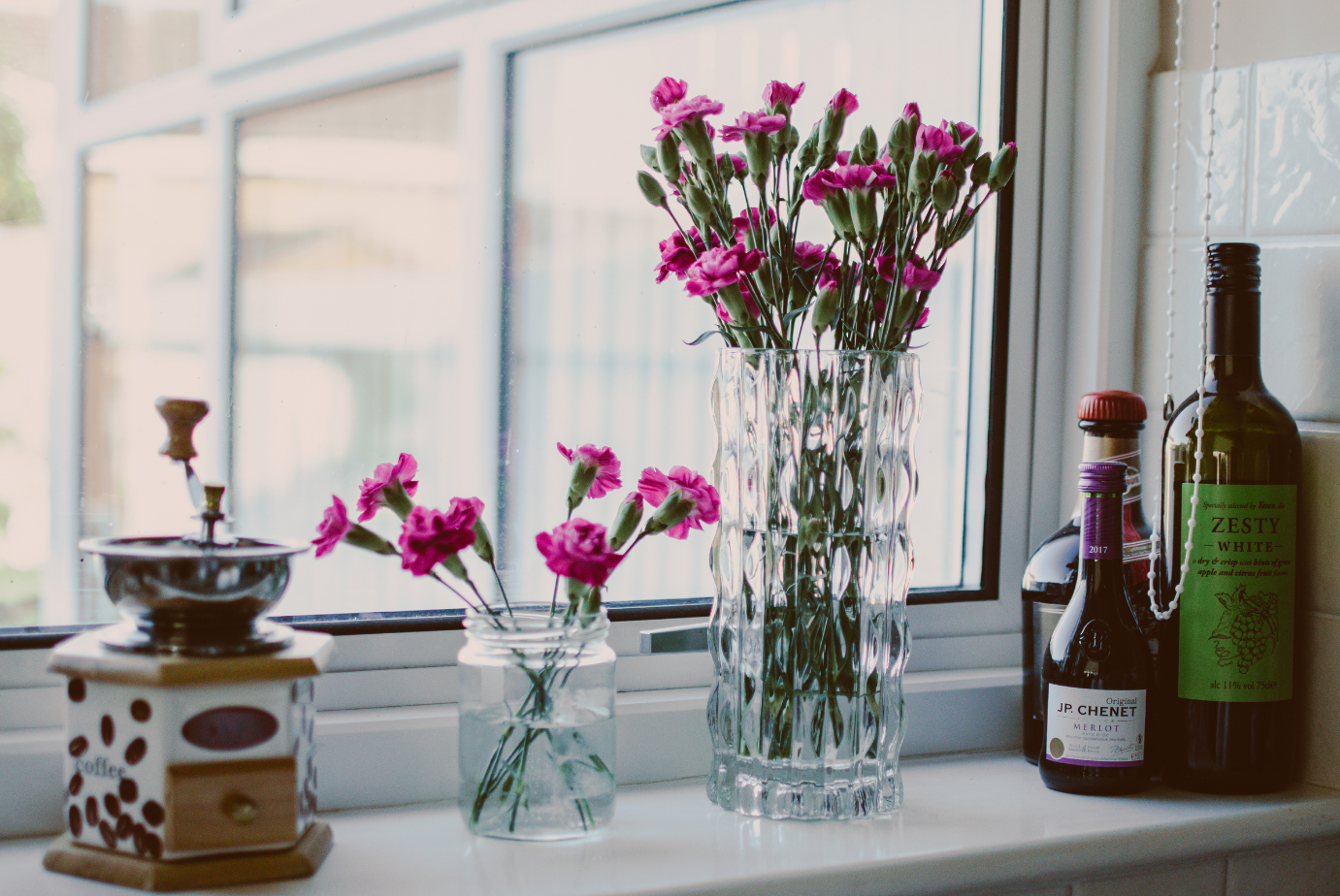 a photograph of a window with a coffee grinder, pink flowers in a vase and wine bottles on the windowsill