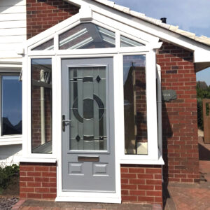 A photograph of a red brick bungalow with a white upvc and brick porch and a grey door with decorative glazing